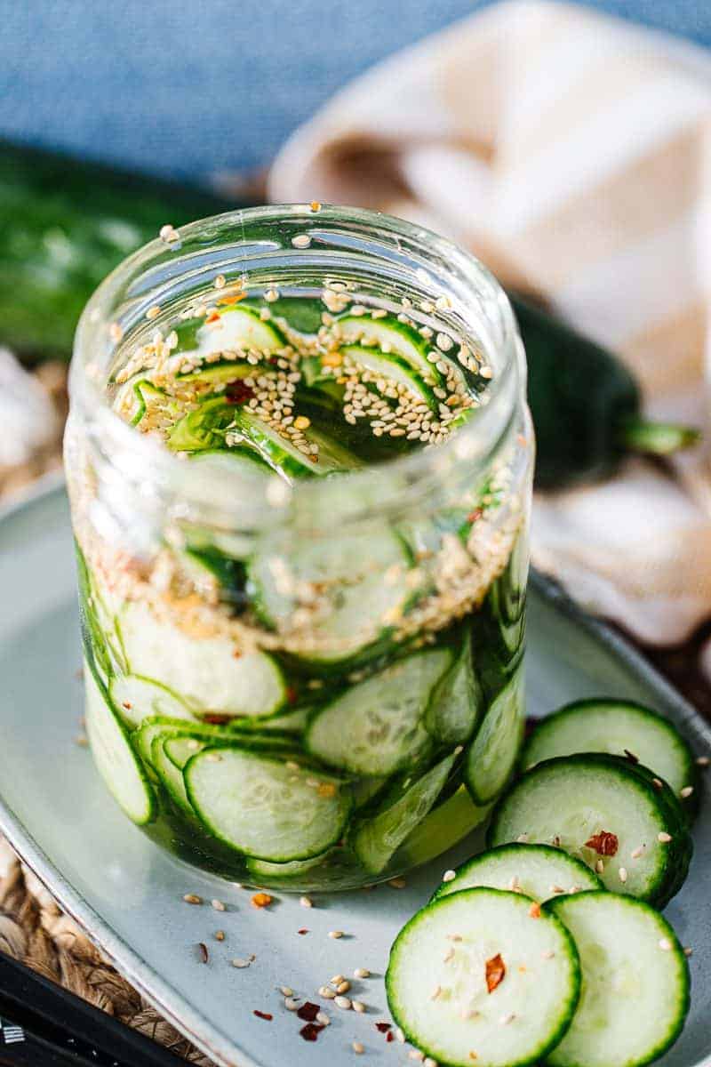 sliced cucumbers with seasonings and liquid in glass jar on an oblong plate next to sliced cucumbers