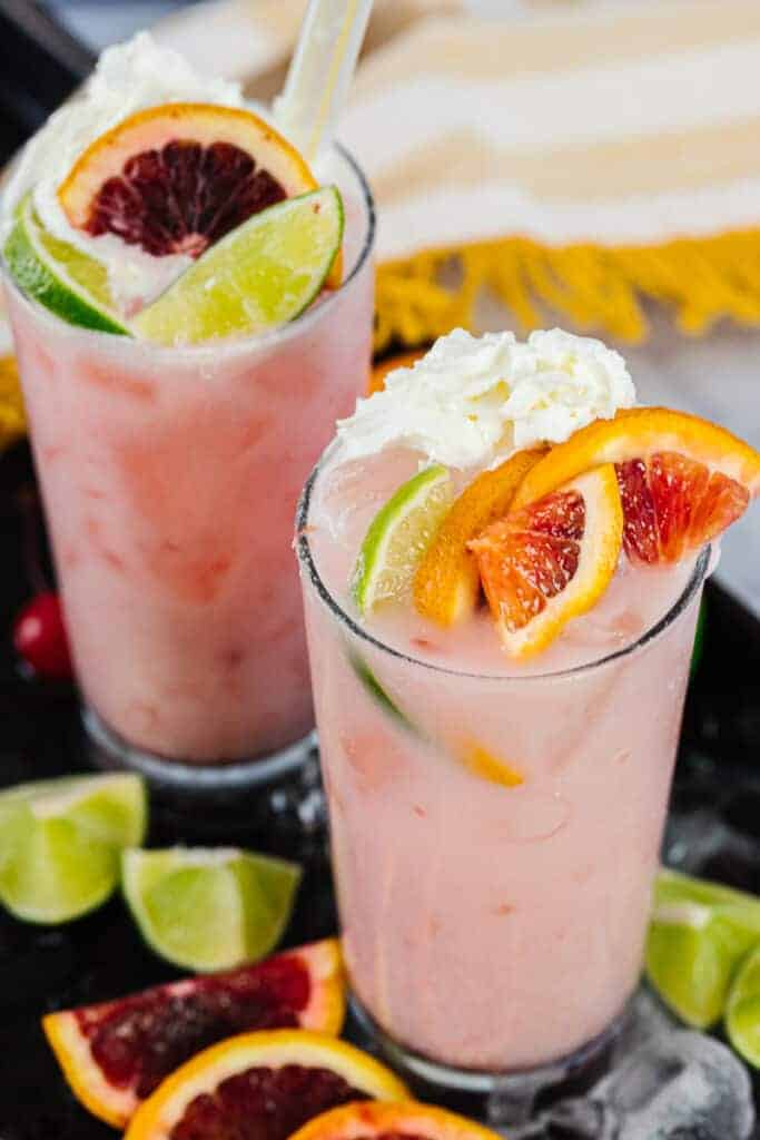 two blood orange coconut cream sodas topped with blood orange slices, lime slices, and whipped cream with ice and on a black tray with ice, lime slices, and blood orange slices in front of a yellow stripped towel