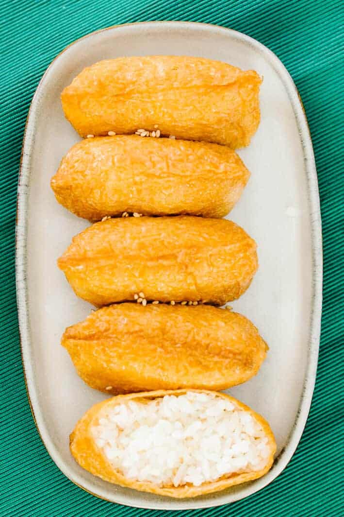 top view of five inari in a vertical row on a white plate and teal cloth with bottom inari open revealing white rice