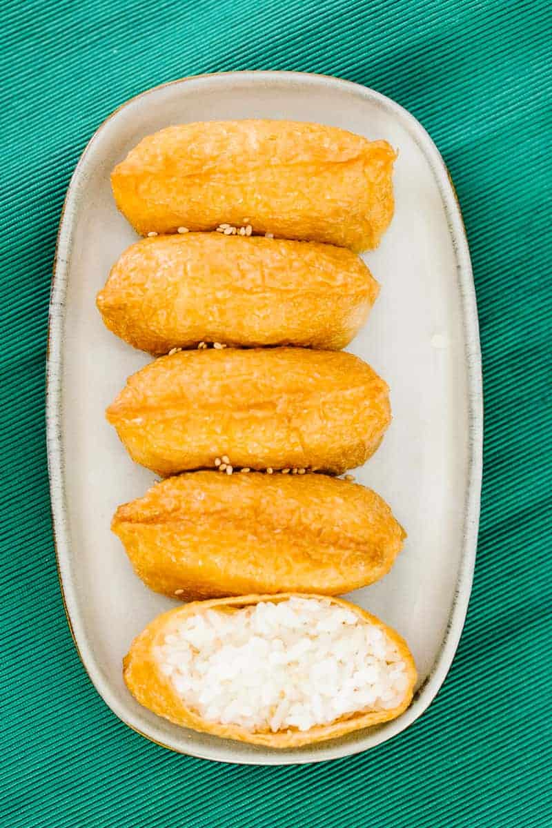 top view of five inari in a vertical row on a white plate and teal cloth with bottom inari open revealing white rice