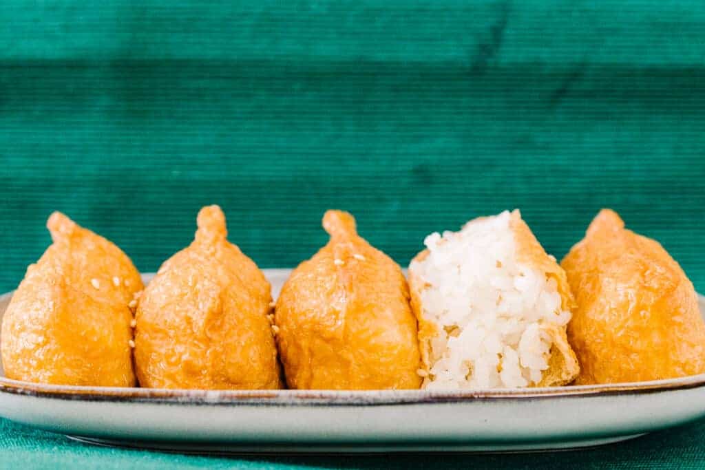 side view of five inari on a cream plate and teal cloth background with second from the right inari open to reveal white rice