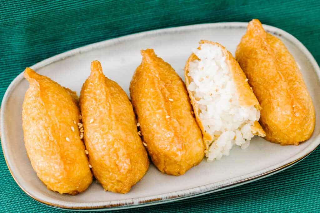 top view of five inari on a cream plate and teal cloth background with second from the right inari open to reveal white rice