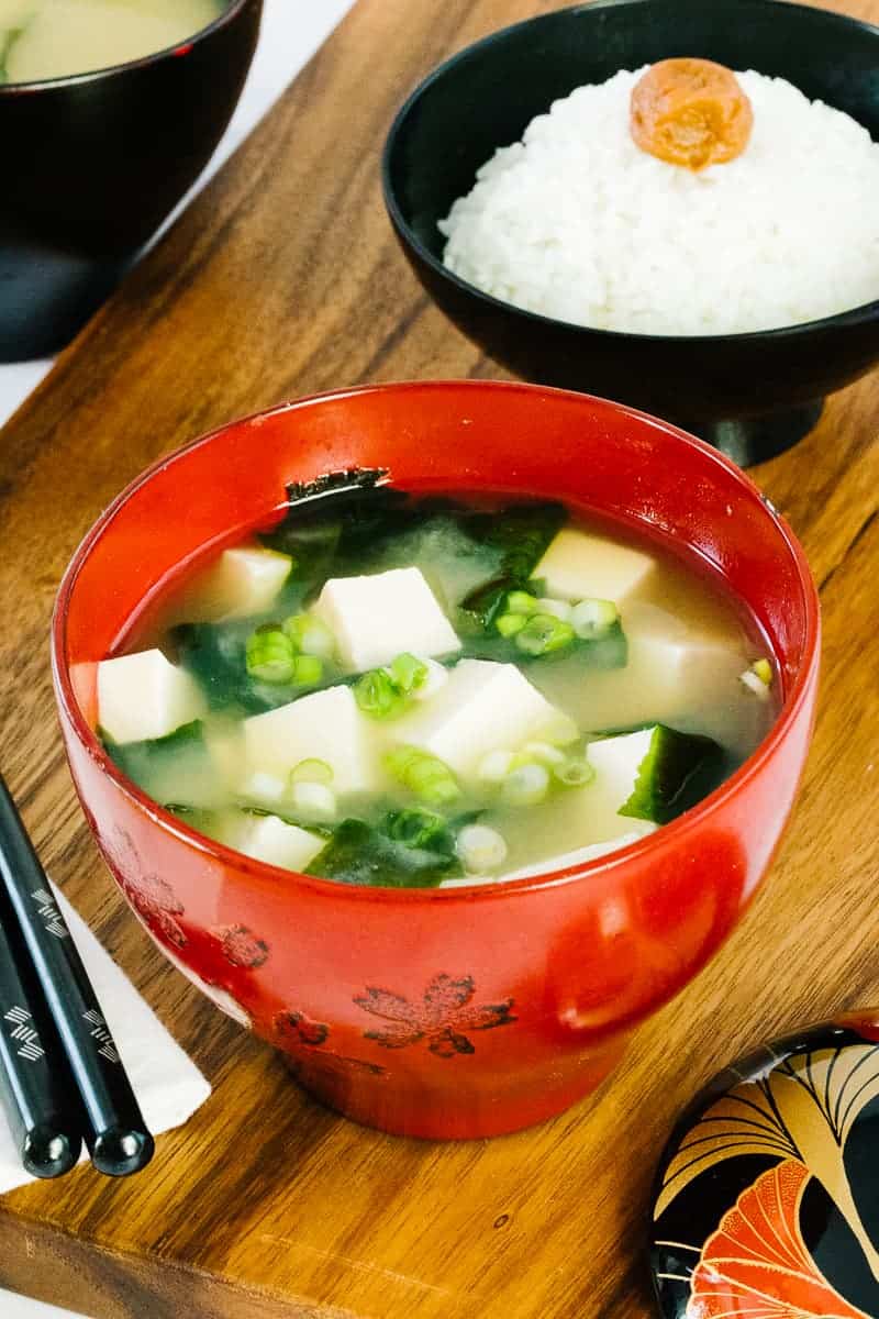 miso soup in red bowl in front of black bowl of white rice next to chopsticks on a wood board