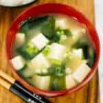 top view of miso soup in red bowl next to chopsticks in front of black bowl of white rice on a wood board