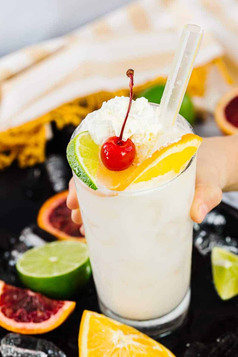 hand reaching out holding simple orange coconut cream soda topped with whipped cream, an orange slice, lime slice and cherry on a black tray with citrus slices in front of yellow striped towel