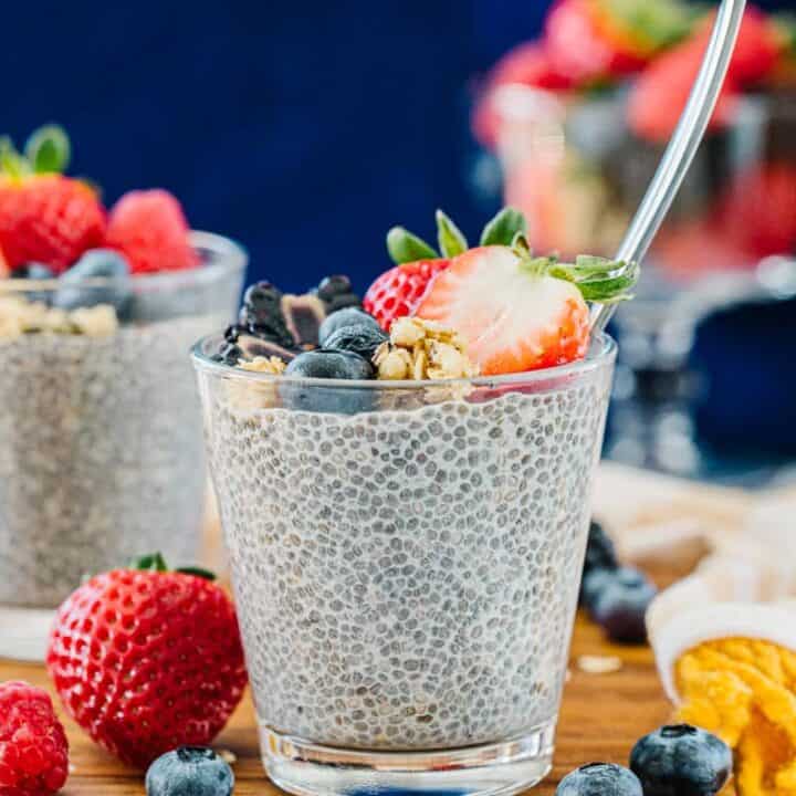 Glass cup of chia pudding with spoon topped with strawberries, blueberries, blackberries, and granola next to blueberries, strawberries, and oats on a wooden board with bowl of strawberries behind