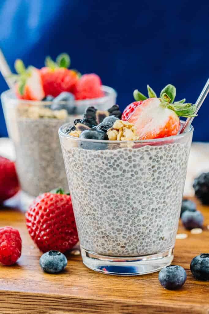 Two glass cups of chia pudding with spoon topped with strawberries, blueberries, blackberries, and granola next to blueberries, strawberries, and oats on a wooden board