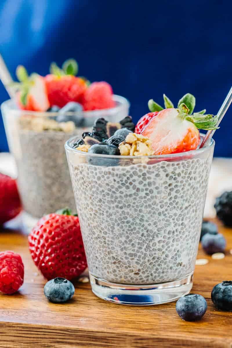 Two glass cups of chia pudding with spoon topped with strawberries, blueberries, blackberries, and granola next to blueberries, strawberries, and oats on a wooden board