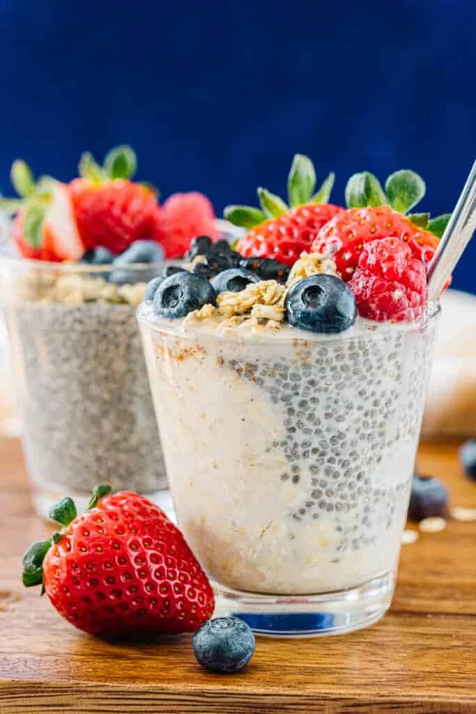 Two glass cups of chia pudding and overnight oats with spoon topped with strawberries, blueberries, blackberries, and granola next to blueberries, strawberries, and oats on a wooden board