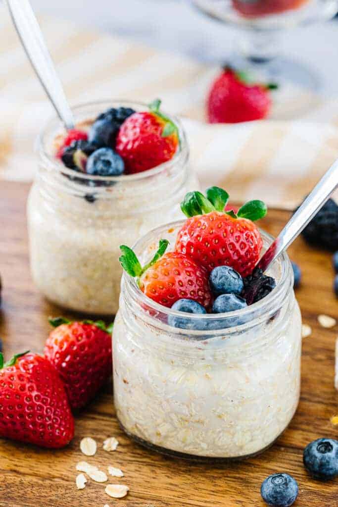 Close up of two glass jars with overnight oats with spoon topped with strawberries, blueberries, and blackberries next to strawberries, blueberries, and oats on a wooden board next to yellow and white striped towel