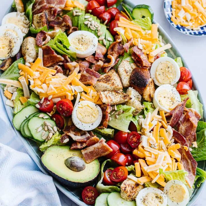 top view of assembled cobb salad surrounded by bowl of cheese, tomatoes, and white cloth