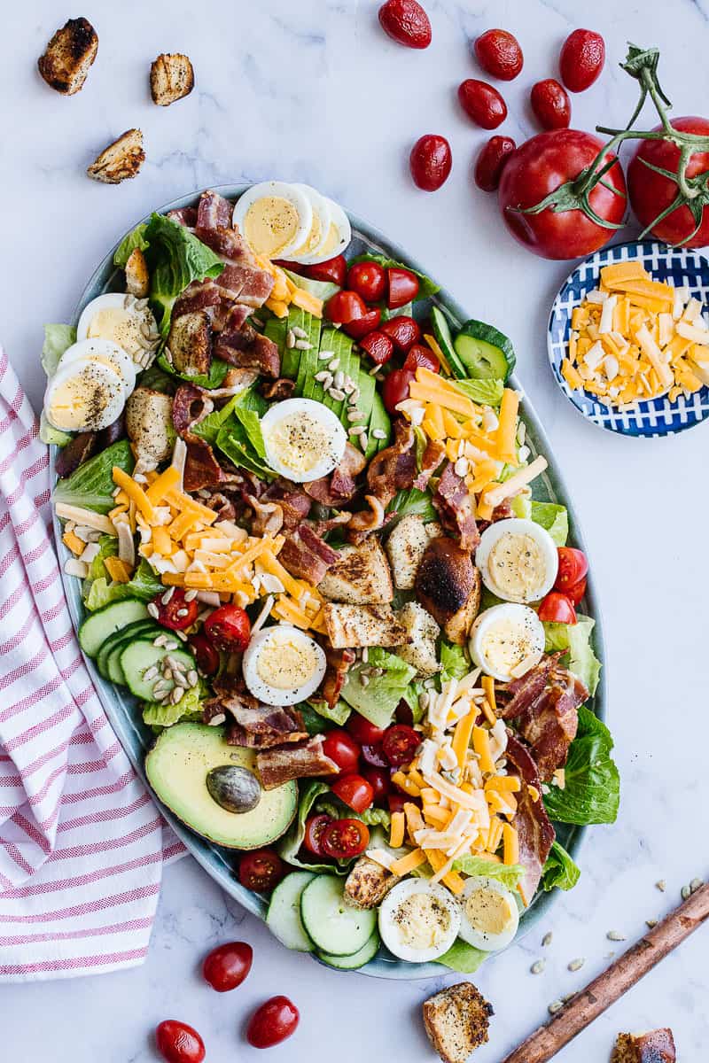 top view of assembled cobb salad surrounded by bowl of cheese, tomatoes, brown bar and striped cloth
