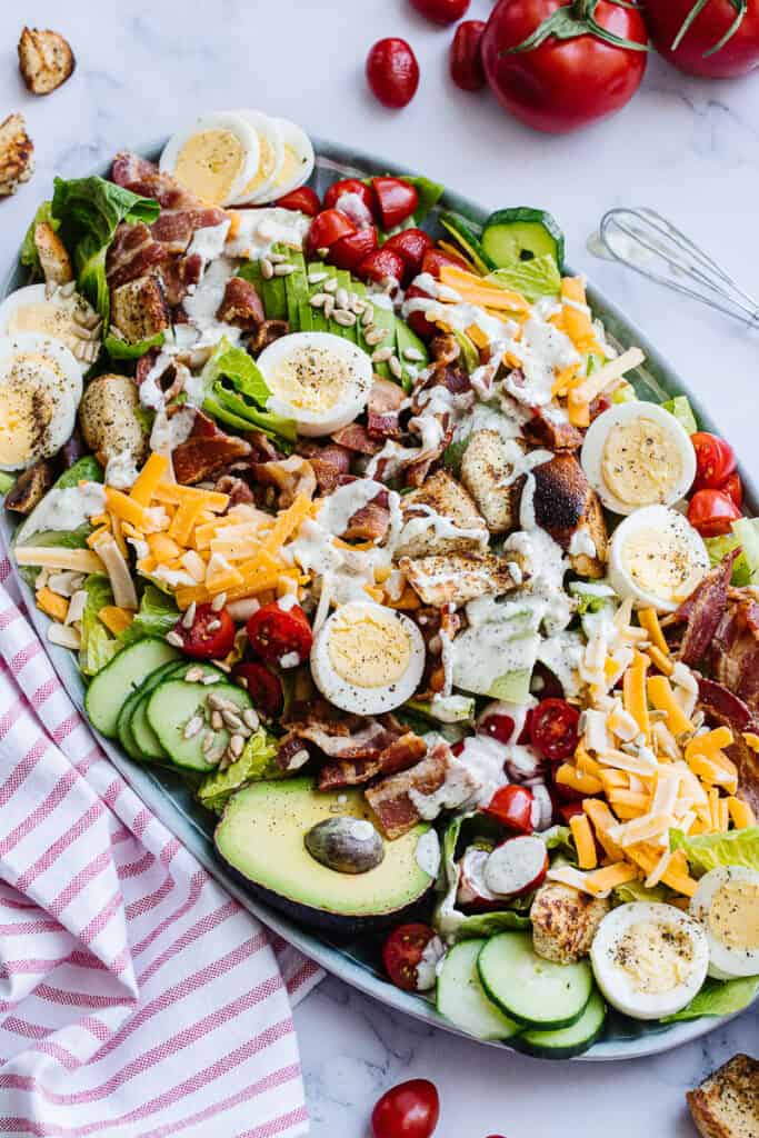 Assembled cobb salad surrounded by tomatoes, whisk, and striped cloth