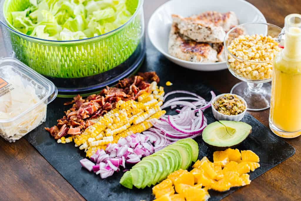 ingredients for catana salad (bacon, corn, red onions, avocados, oranges) sliced on black cutting board surrounded by bowls of lettuce, chicken, corn, cheese, and vinaigrette