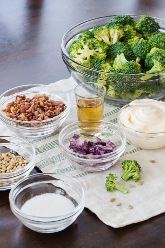 broccoli salad ingredients placed in glass bowls on a white cloth on a dark table