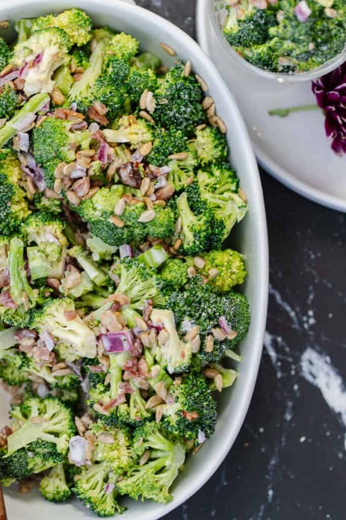 close up top view of broccoli salad in white bowl next to white plate with smaller bowl of broccoli salad and a purple flower