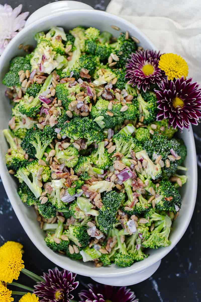 top view of broccoli salad in white serving bowl with purple and yellow flowers next to it on a dark surface next to white cloth and more flowers