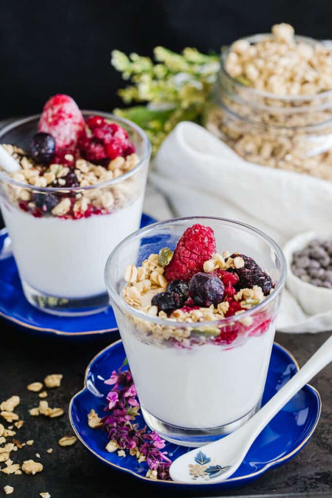two glass cups of instant pot yogurt topped with granola, seeds, and berries in front of a white cloth topped with foliage and a jar of granola