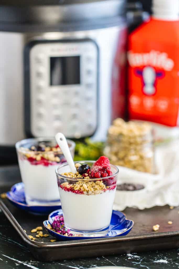 two cups of instant pot yogurt topped with granola and berries in front of instant pot, fairlife milk, and jar of granola