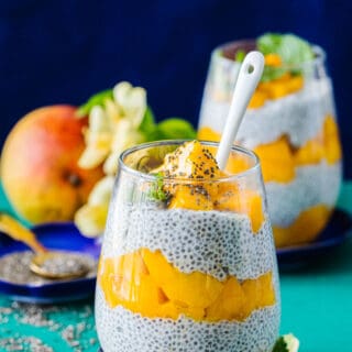 side view of coconut mango chia pudding topped with mangos and chia seeds on a blue plate with another glass of pudding and a mango behind