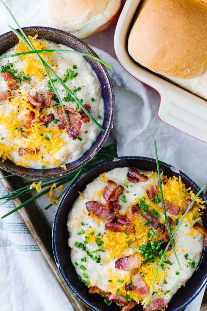 top view of two bowls of loaded baked potato soup in dark bowls next to a pan of rolls on a white cloth