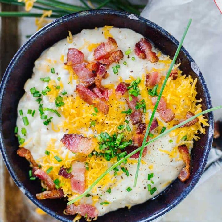 close up top view of loaded baked potato soup in a dark bowl on a white cloth and pan next to a spoon
