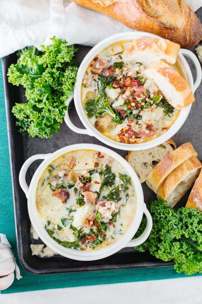 top view of two bowls of zuppa toscana soup in white bowls next to bread and greens on a black board on a teal cloth