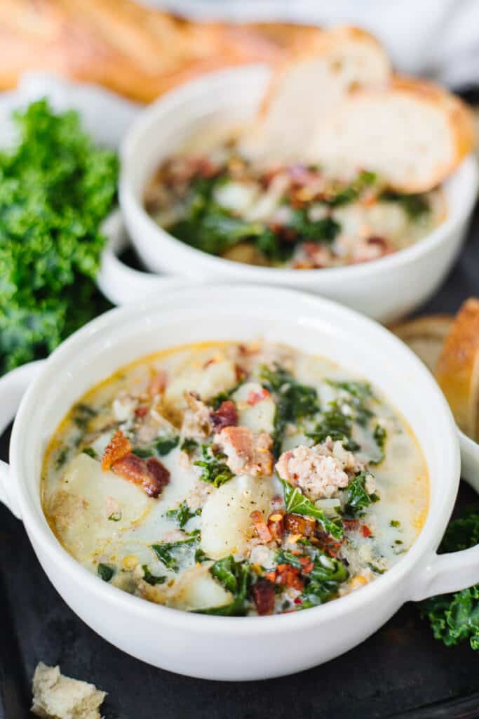 close up of zuppa toscana soup in a white bowl in front of another bowl next to bread and greens on a black board