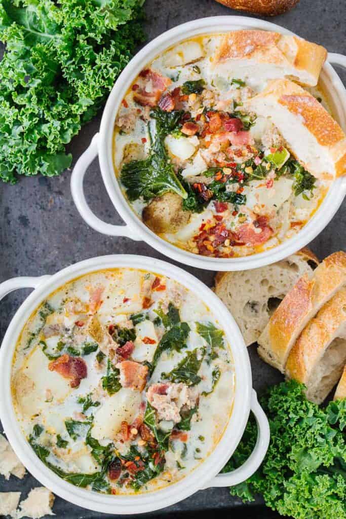 top view of two bowls of zuppa toscana soup in white bowls next to bread and greens on a black board