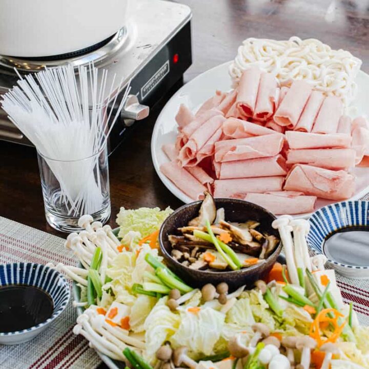 plate of shabu shabu vegetables on plate in front of sliced pork rolls and noodles on a plate next to pot of water and bowls of soy sauce