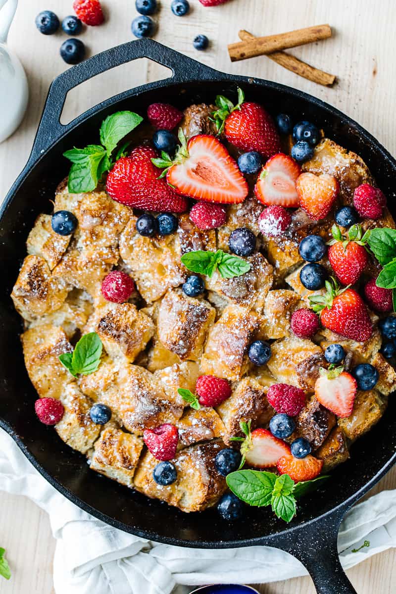 Top view of bread pudding mixed with strawberries, blueberries, raspberries, and greenery in a black cast iron pan