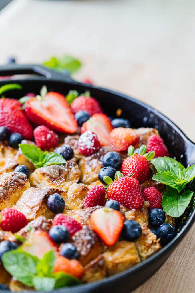 close up of bread pudding mixed with strawberries, blueberries, raspberries, and greenery in a black cast iron pan