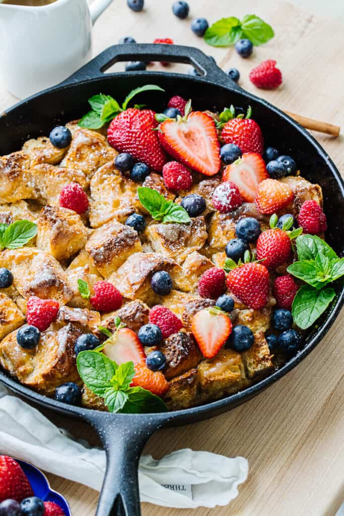 side view of bread pudding mixed with strawberries, blueberries, raspberries, and greenery in a black cast iron pan.