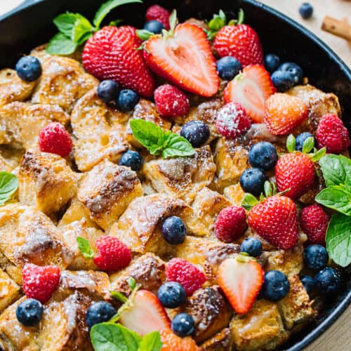 close up of bread pudding mixed with strawberries, blueberries, raspberries, and greenery in a black cast iron pan.