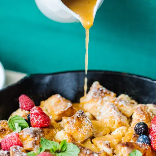 pitcher of syrup being poured on bread pudding, strawberries, raspberries, and blueberries in a cast iron skillet