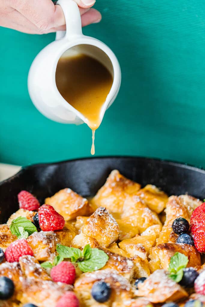 pitcher of syrup being poured on bread pudding, strawberries, raspberries, and blueberries in a cast iron skillet