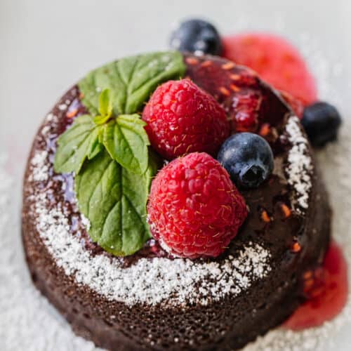 close up of round chocolate cake dusted with powdered sugar next and topped with berries and syrup