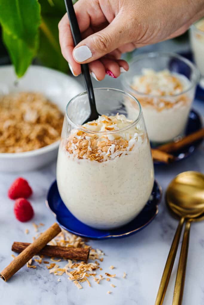hand with spoon mixing coconut rice pudding topped with shredded toasted coconuts in a clear glass on a blue plate next to cinnamon sticks, gold spoons, and bowl of toasted coconut