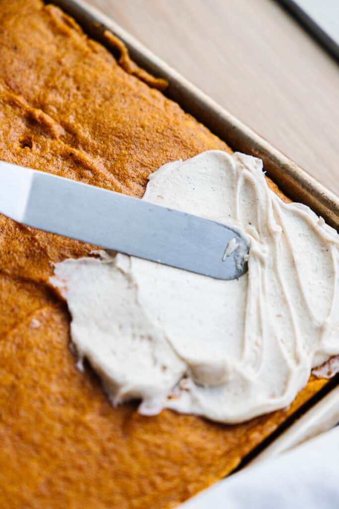 knife spreading cream cheese on baked pumpkin bars in a sheet pan